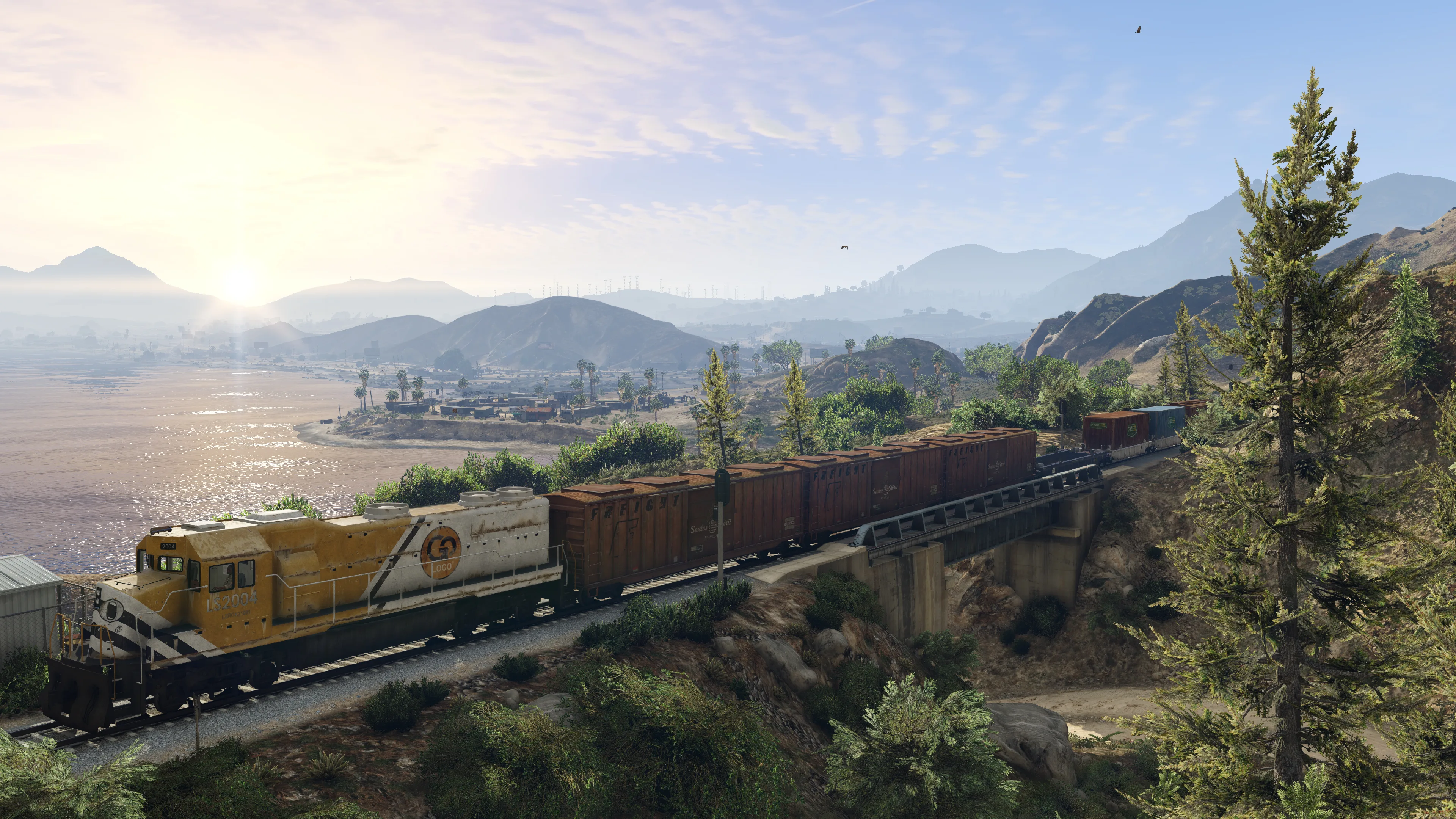A freight train crosses a bridge through hills beside a large body of water in early morning light.