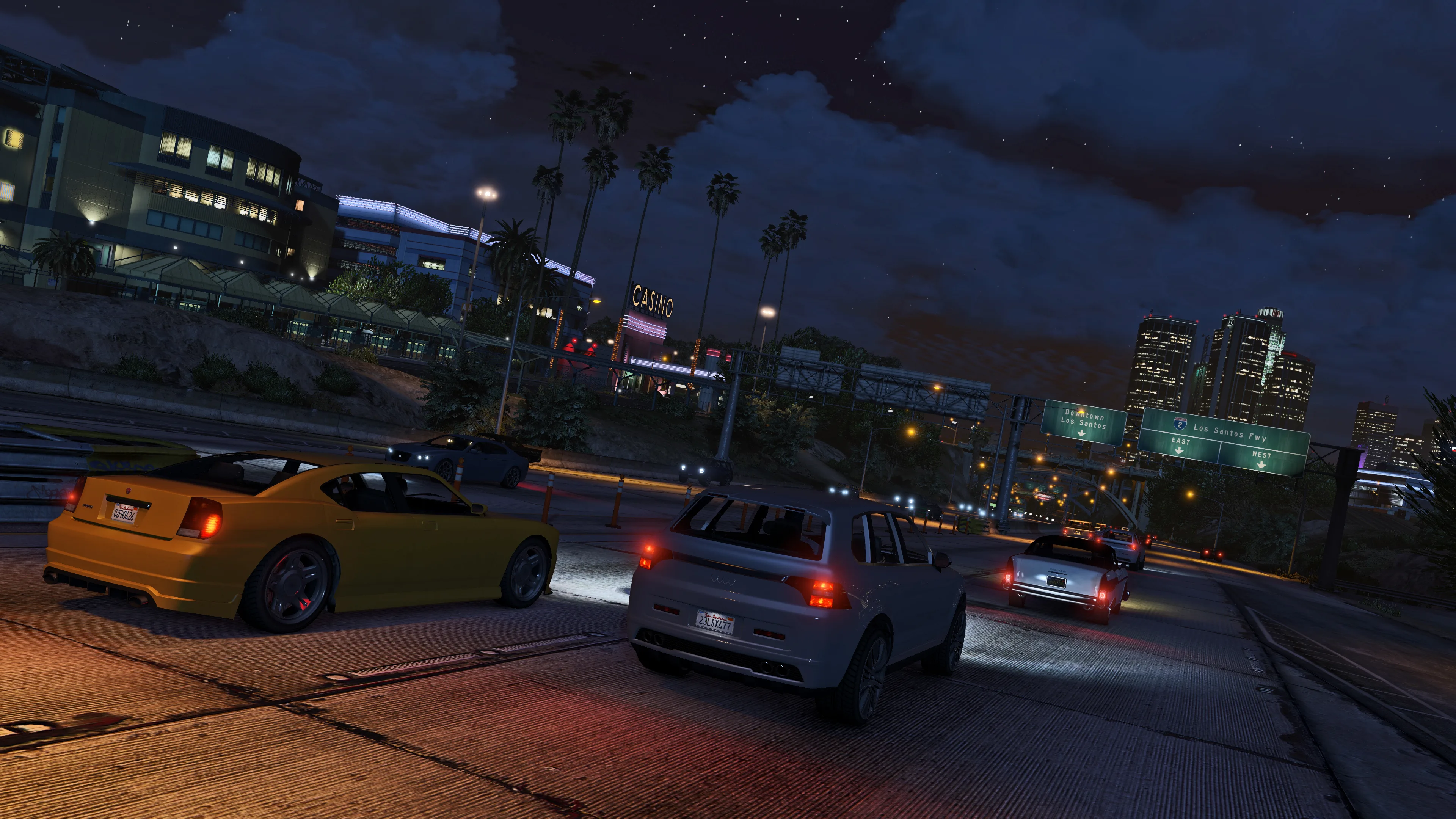 Cars travel along a Los Santos freeway at night beneath city lights and road signs.